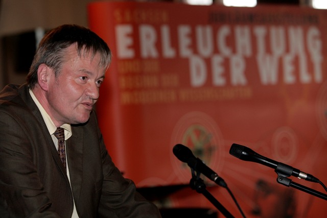 Ein Mann steht an einem Rednerpult und spricht in Mikrofone. Im Hintergrund sieht man einen orange-farbenen Aufsteller mit der Aufschrift "Erleuchtung der Welt".