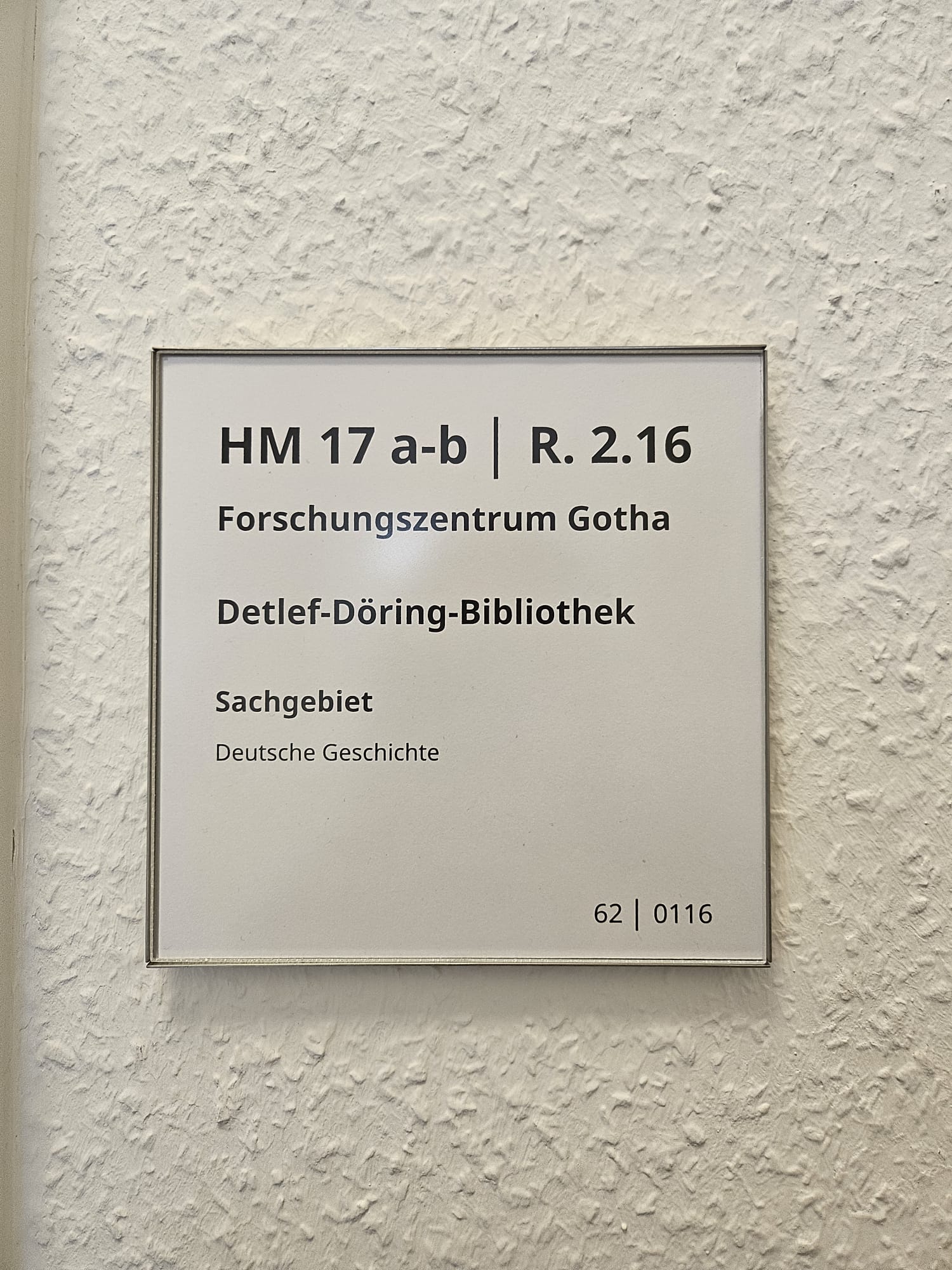 Weißes Beschriftungsschild an einer Wand. Auf dem Schild steht "HM 17 a-b, R 2.16, Forschungsbibliothek Gotha, Detlef-Döring-Bibliothek, Sachgebiet: Deutsche Geschichte, 62, 0116".