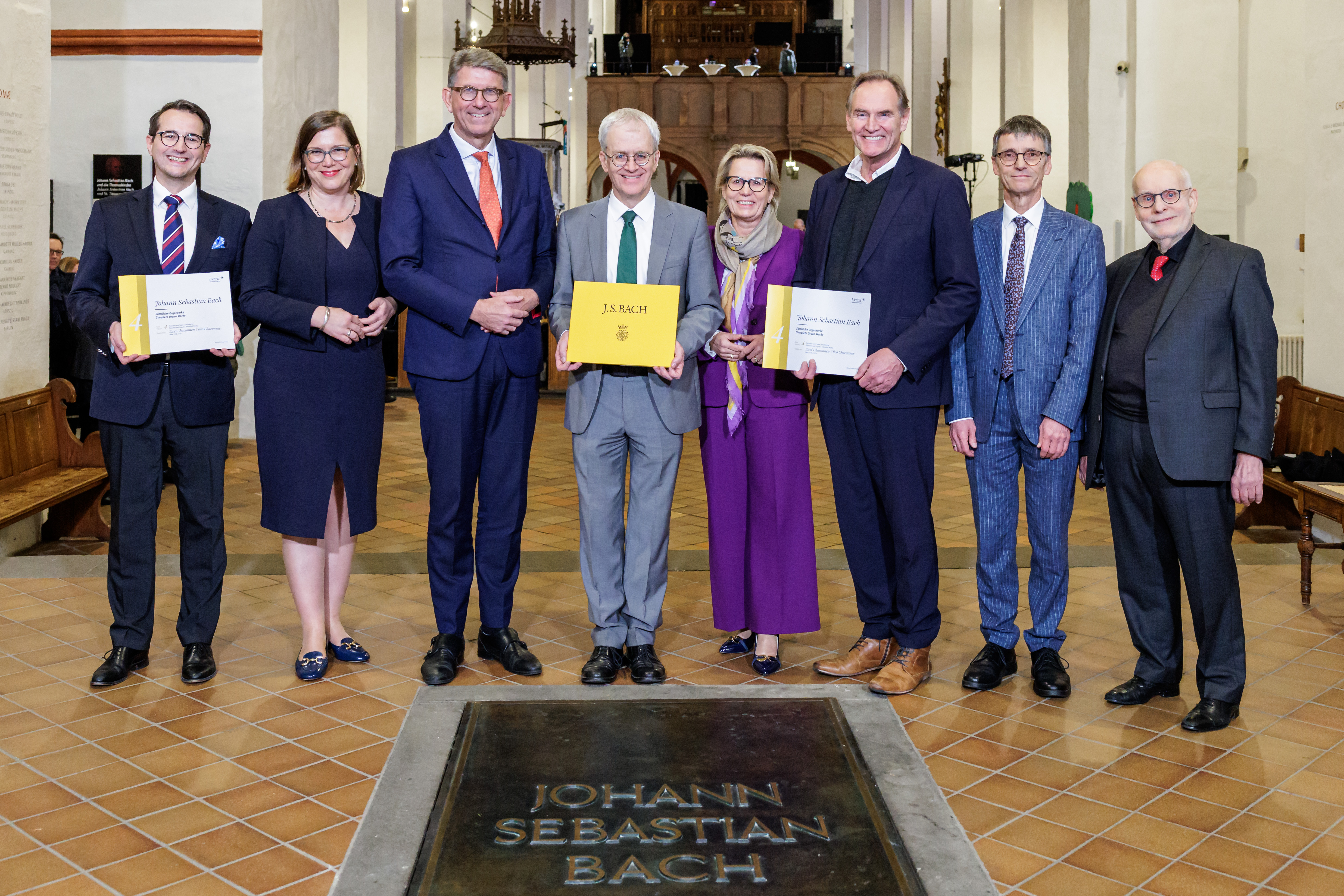 Sechs männliche und zwei weibliche Personen stehen hinter der Grabplatte Johann Sebastian Bachs in der Thomaskirche Leipzig. Drei von ihnen halten gelbe Notenbände in den Händen.
