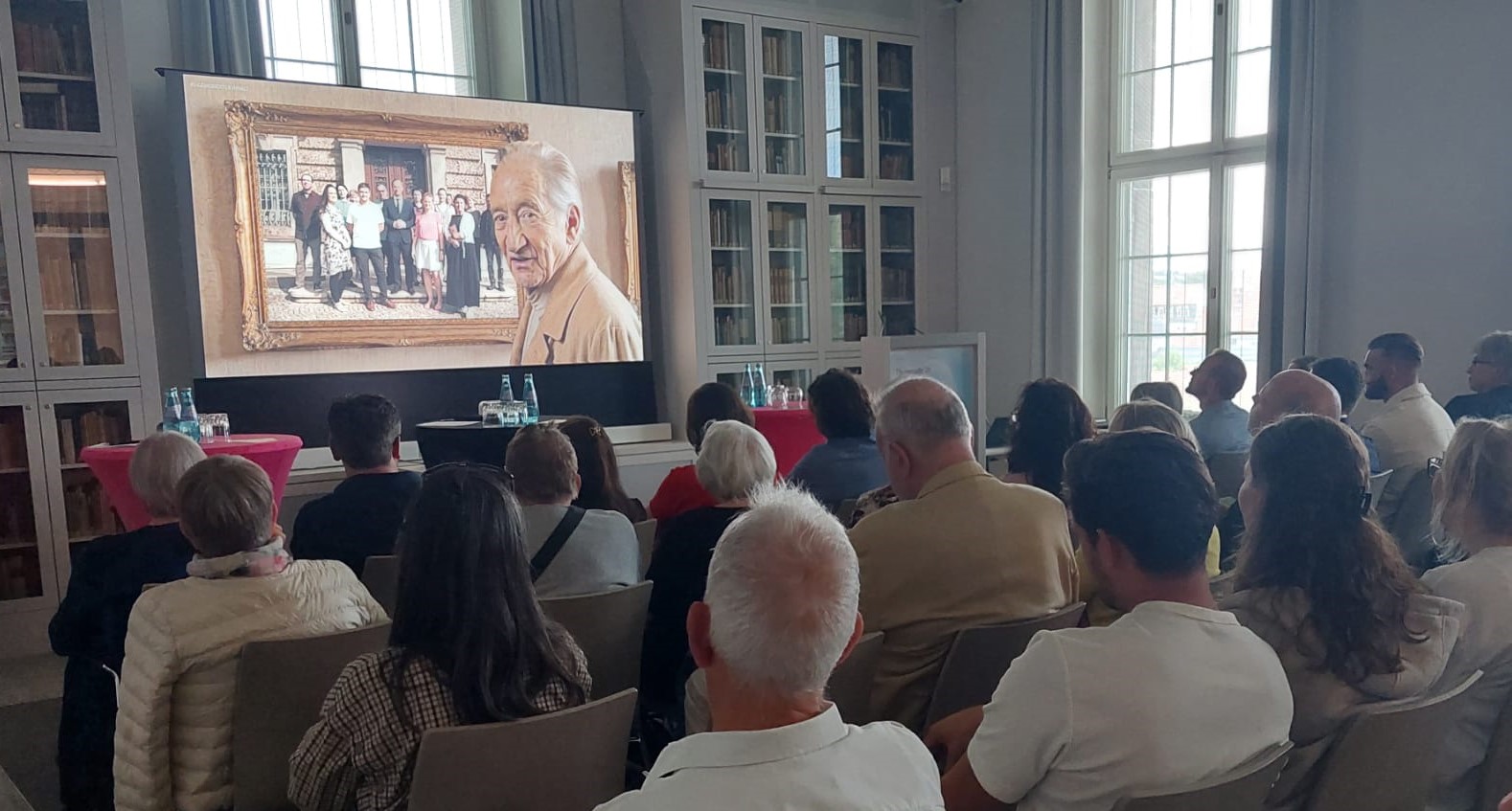Eine Gruppe von Menschen sitzt auf Stühlen in einem historischen Vortragssaal mit Büchervitrinen an den Wänden. Sie schauen auf einen Monitor, auf dem ein KI-generierter Goethe zu sehen ist, der auf ein Bild mit Mitarbeitern des Goethe- und Schiller-Archivs blickt.