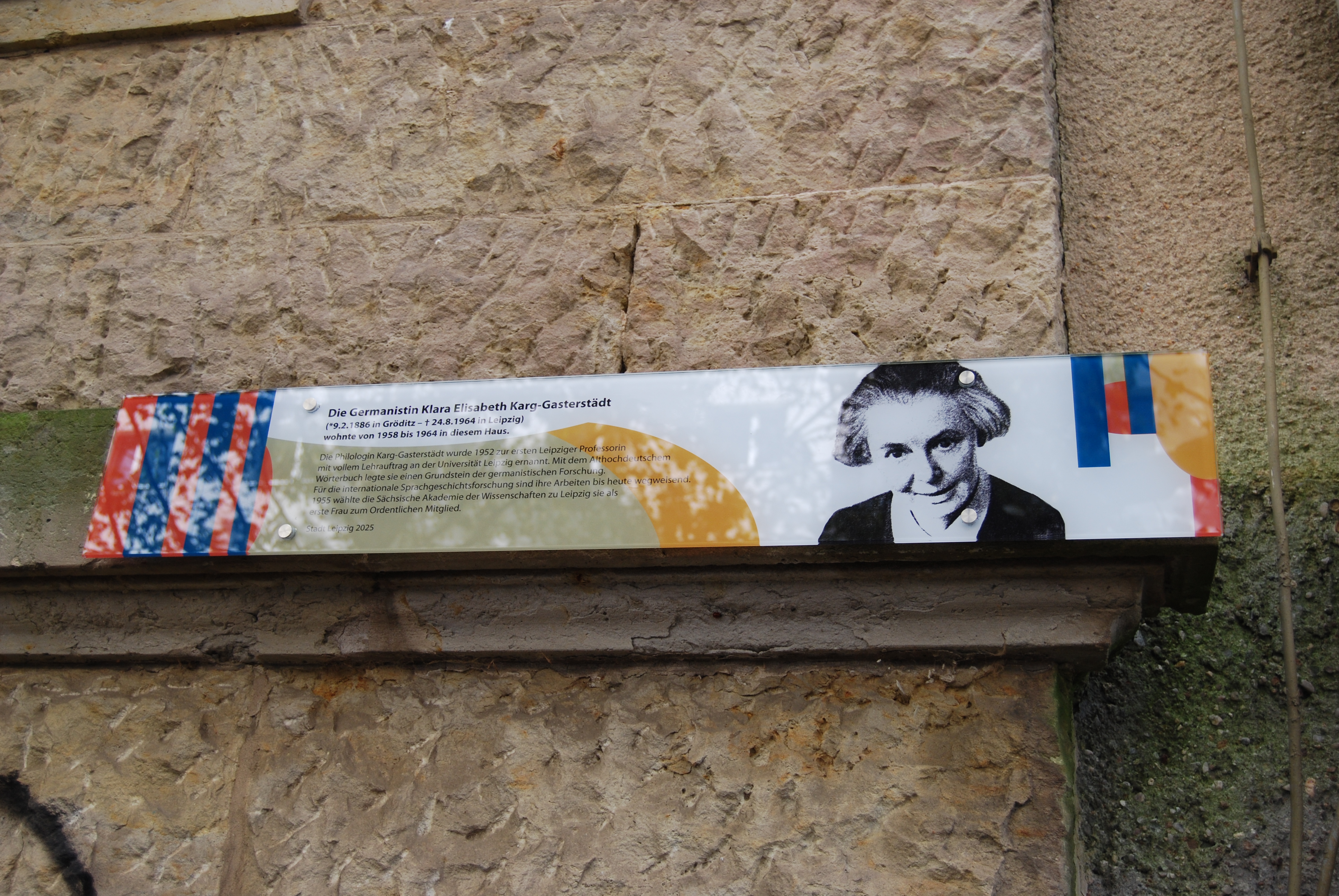 Gedenkplakette an einer Hausmauer. Man sieht einen Text über Elisabeth Karg-Gasterstädt und ihr Porträtbild. Das ganze wird umrahmt von verschiedenen grafischen Farbelementen.