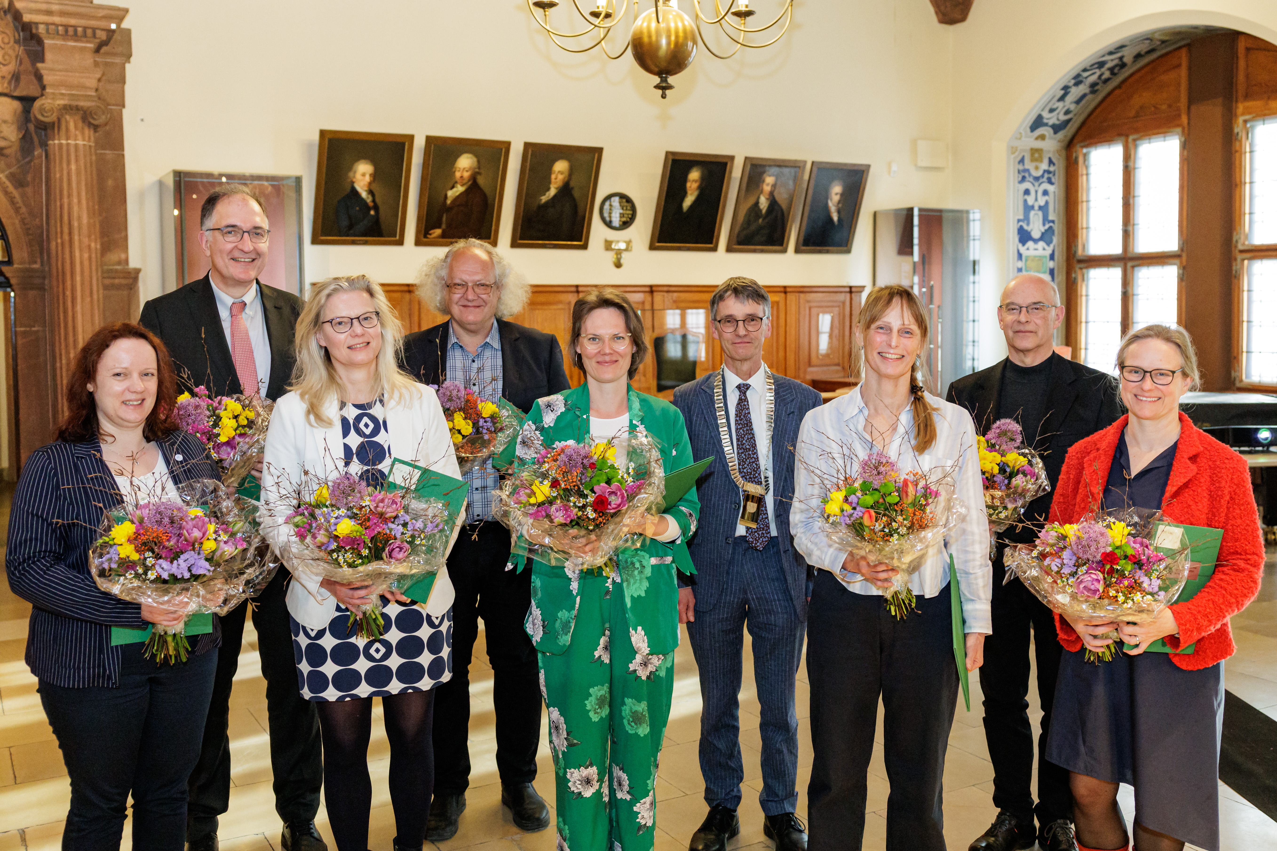 Eine Gruppe von fünf weiblichen und vier männlichen Personen steht lächelnd in einem Festsaal. Im Hintergrund sieht man Gemälde an den Wänden, oben einen Kronleuchter. Die männliche Person in der Mitte trägt eine goldene Amtskette. Die anderen Personen halten Blumensträuße und grüne Urkunden in den Händen.