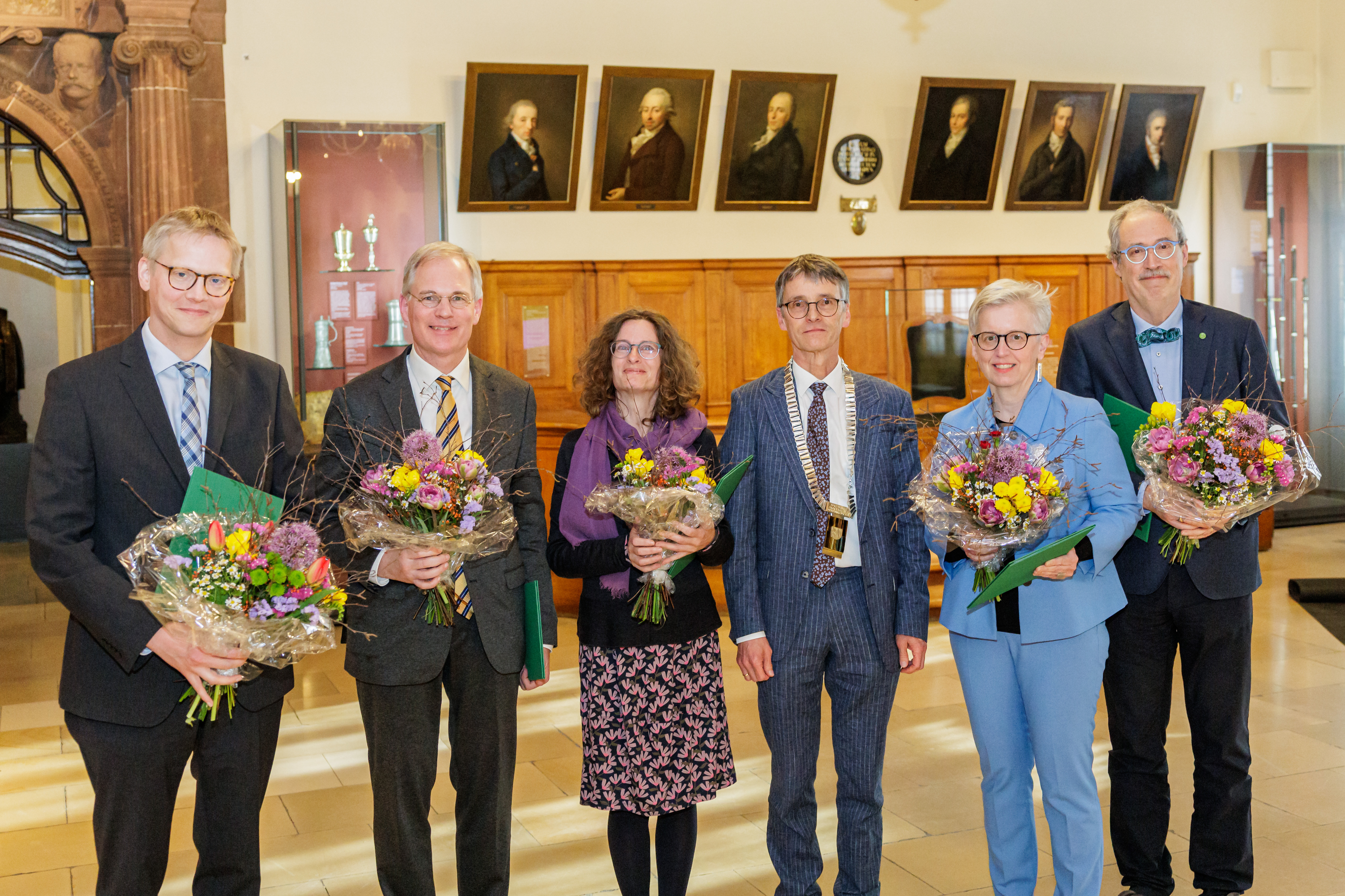 Eine Gruppe von zwei weiblichen und vier männlichen Personen steht lächelnd in einem Festsaal. Im Hintergrund sieht man Gemälde an den Wänden. Die männliche Person in der Mitte trägt eine goldene Amtskette. Die anderen Personen halten Blumensträuße und grüne Urkunden in den Händen.