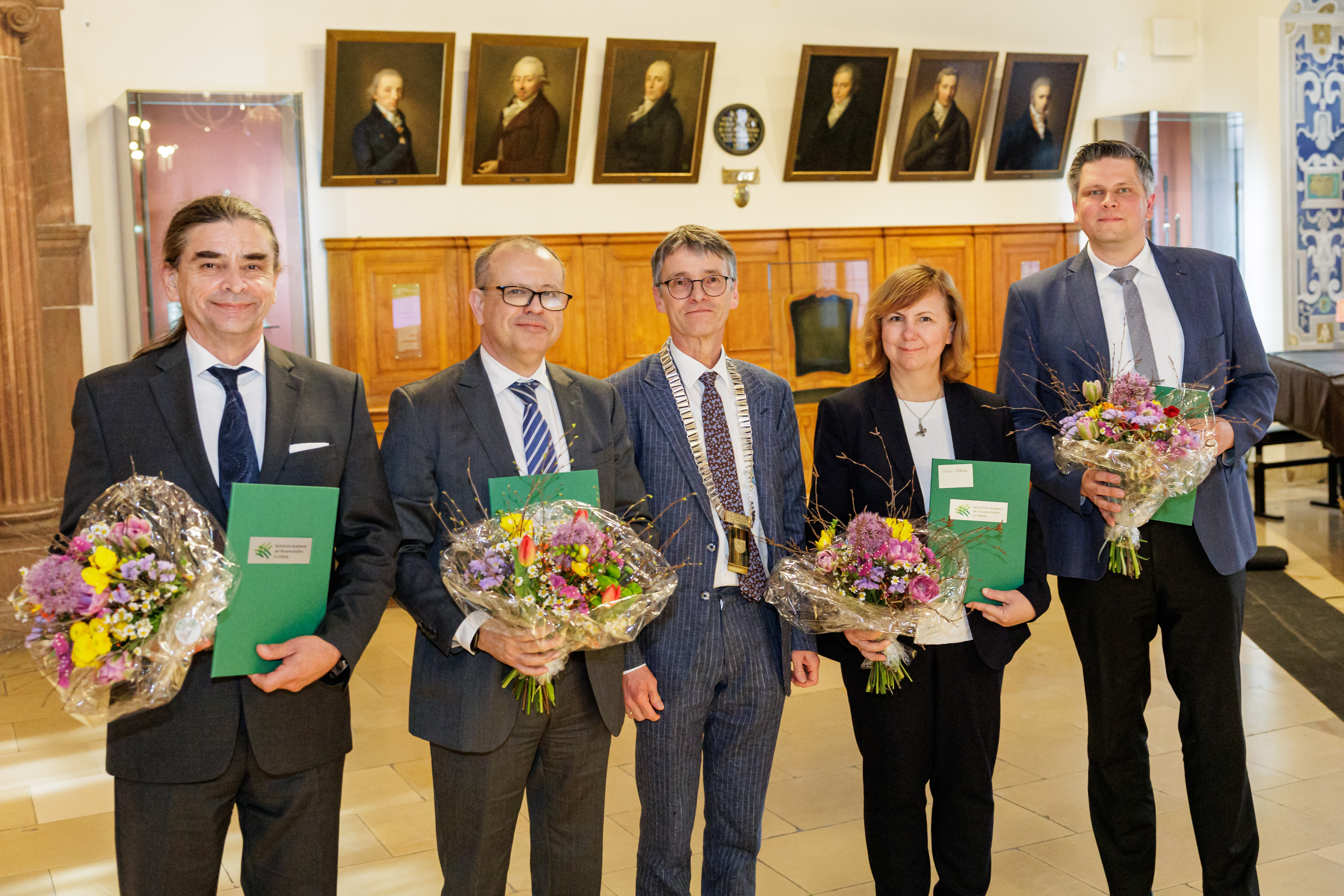 Eine Gruppe von einer weiblichen und vier männlichen Personen steht lächelnd in einem Festsaal. Im Hintergrund sieht man Gemälde an den Wänden. Die männliche Person in der Mitte trägt eine goldene Amtskette. Die anderen Personen halten Blumensträuße und grüne Urkunden in den Händen.