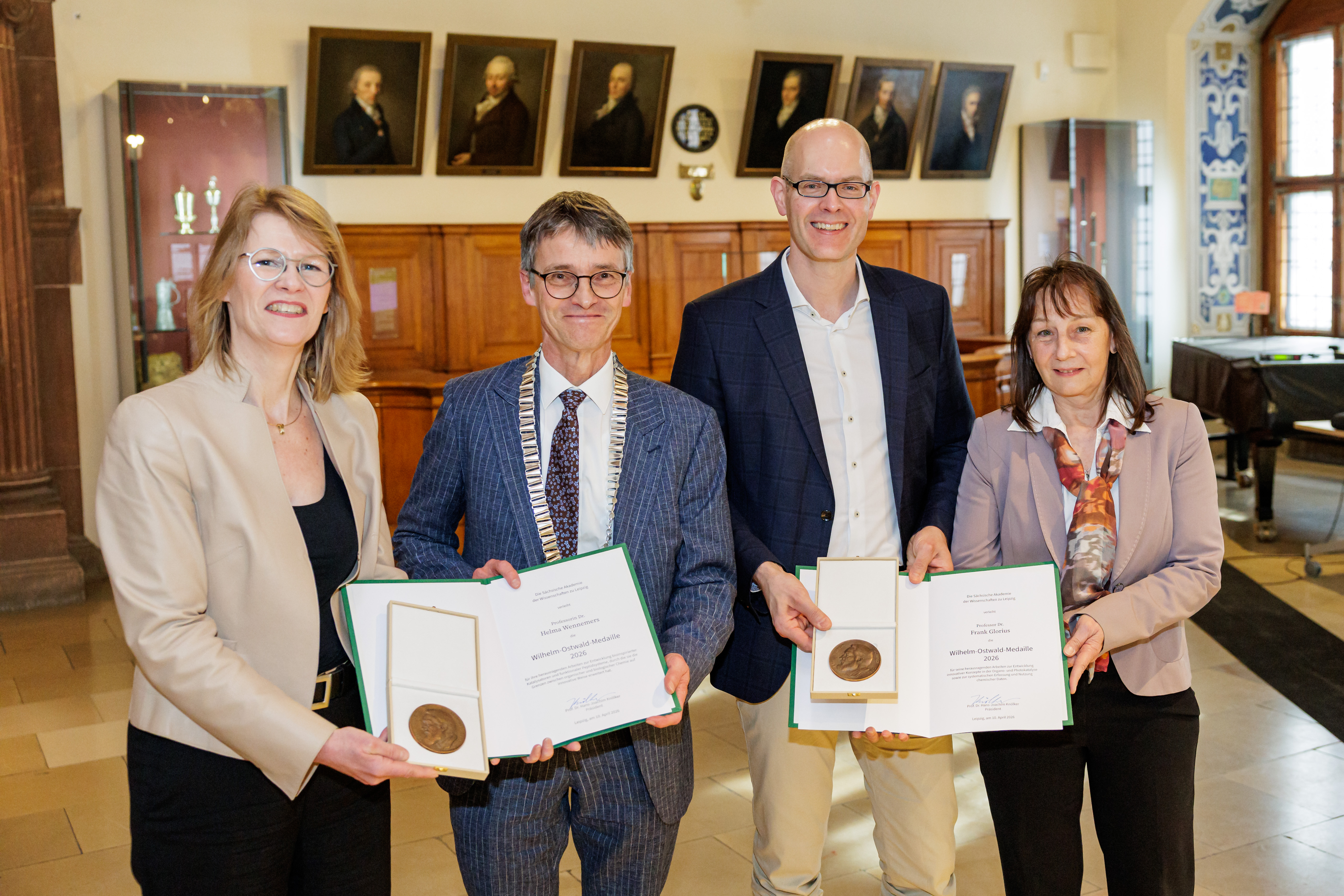 Zwei weibliche und zwei männliche Personen stehen lächelnd in einem Festsaal, gemeinsam halten sie zwei Urkunden und zwei Medaillen in Etuis in den Händen. Im Hintergrund sieht man Gemälde. Eine männliche Person trägt eine goldene Amtskette.