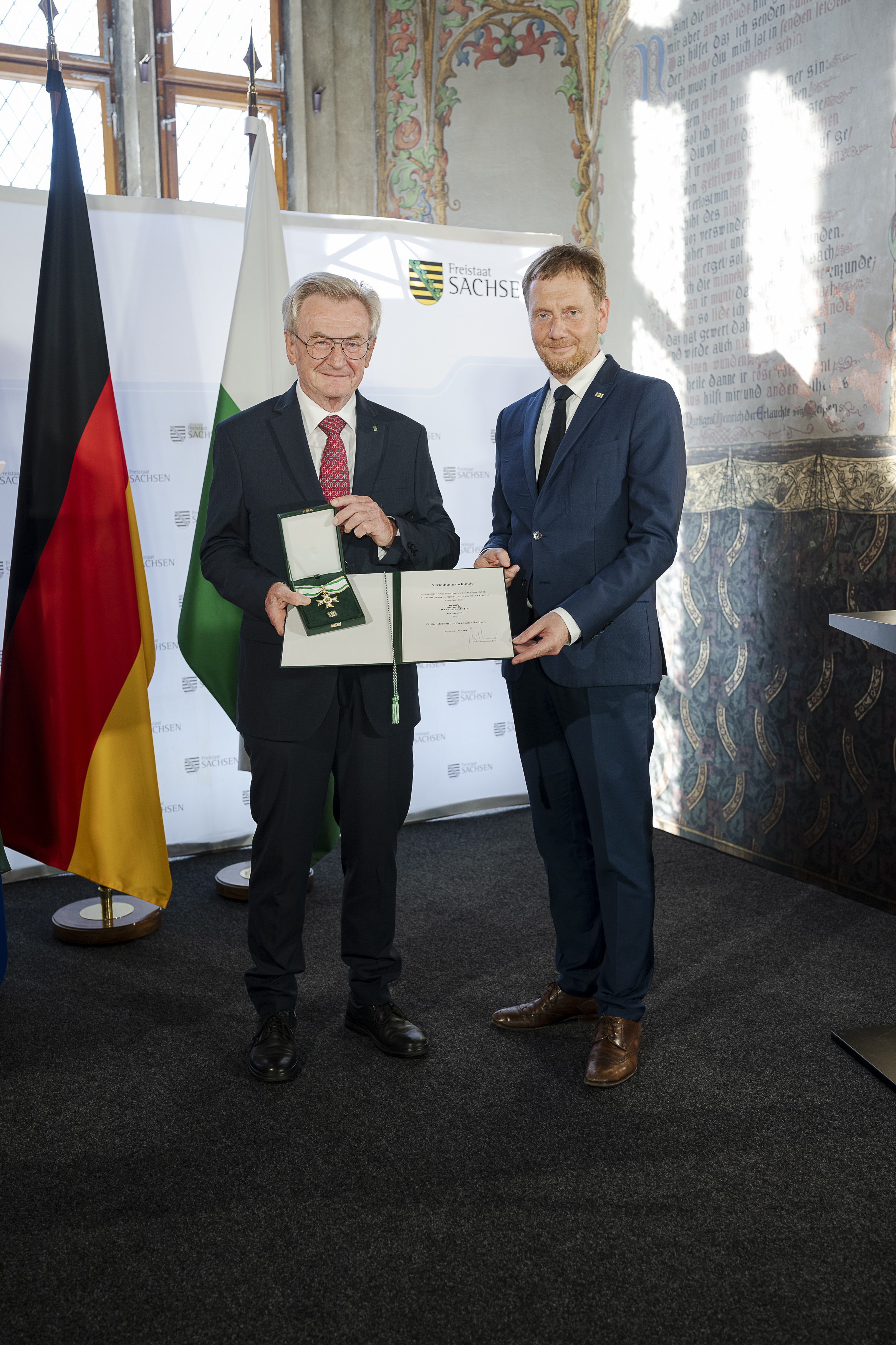 Zwei männliche Personen stehen in einem historischen Saal mit Wandbemalungen vor einer Deutschlandfahne und Aufstellern des Freistaates Sachsen. Sie halten gemeinsam eine Urkunde und einen Orden in die Kamera, beide lächeln.