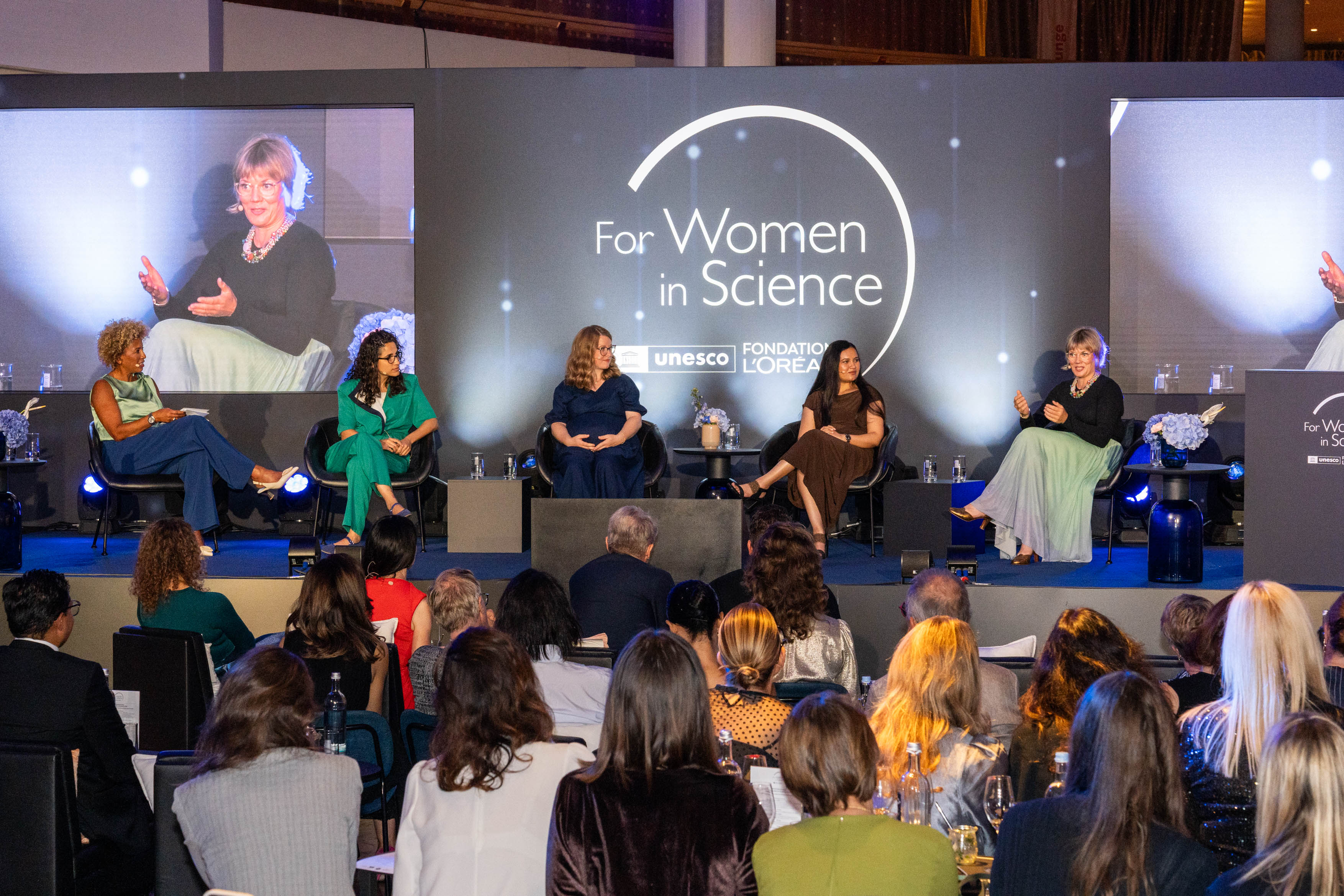 Auf einer Bühne sitzen fünf weibliche Personen mit Headset und diskutieren. Im Hintergrund ist eine Leinwand, auf der "For Women in Science" zu lesen ist.