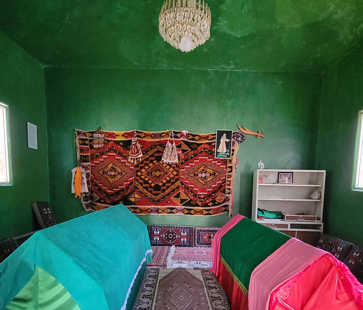 Innenraum eines Mausolems, grüne Wände, Teppiche auf dem Boden und ein Teppich an der Wand, zwei Särge sind mit bunten Tüchern verhüllt.