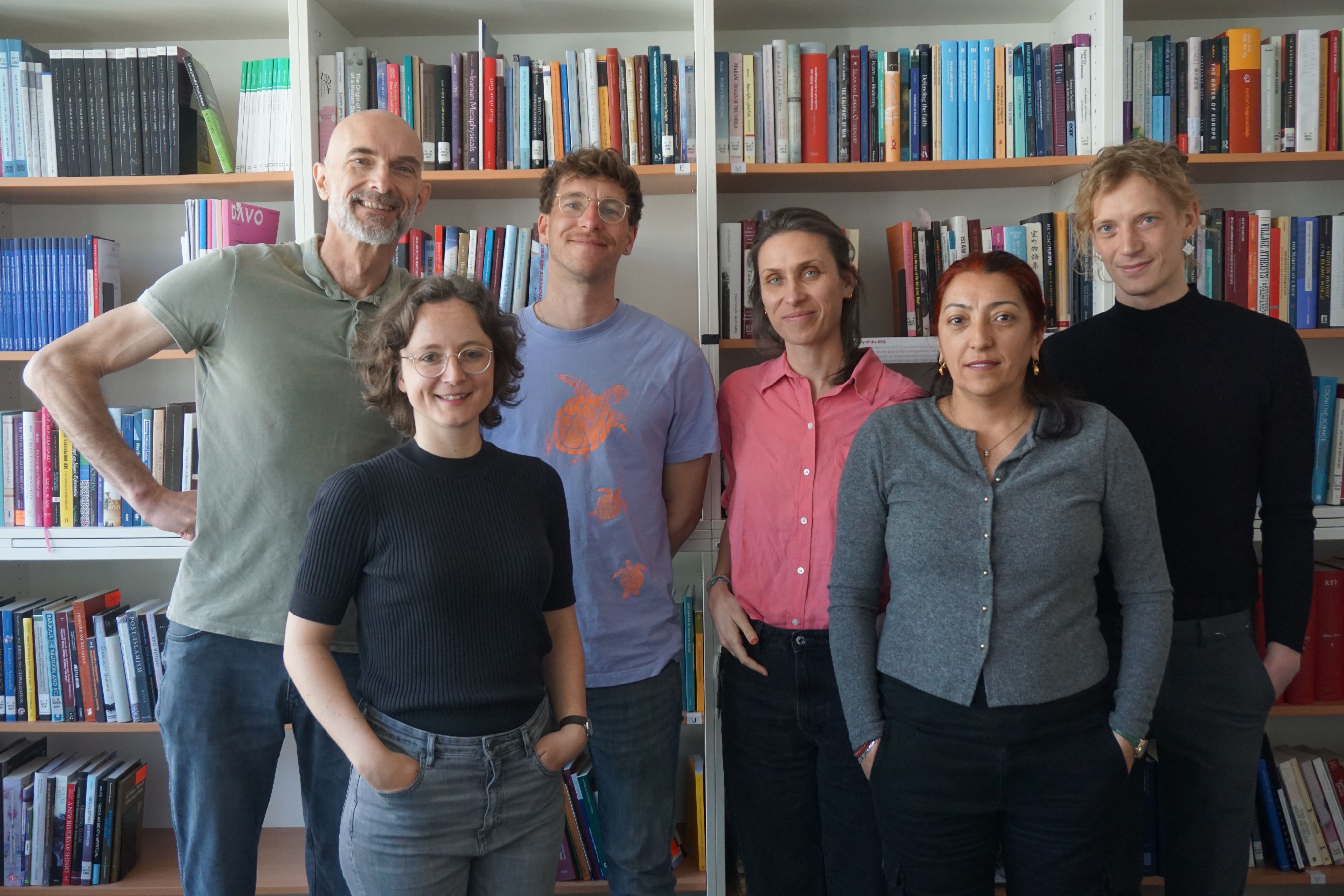 Eine Gruppe von sechs lächelnden Personen steht vor gefüllten Bücherregalen.