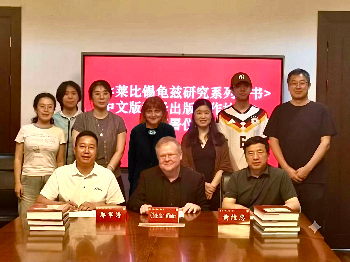 Gruppenfoto mit zehn Personen am Konferenztisch, auf dem Stapel von Büchern liegen. Im Hintergrund ist teilweise ein Flachbildschirm mit weißer Schrift auf rotem Hintergrund zu sehen