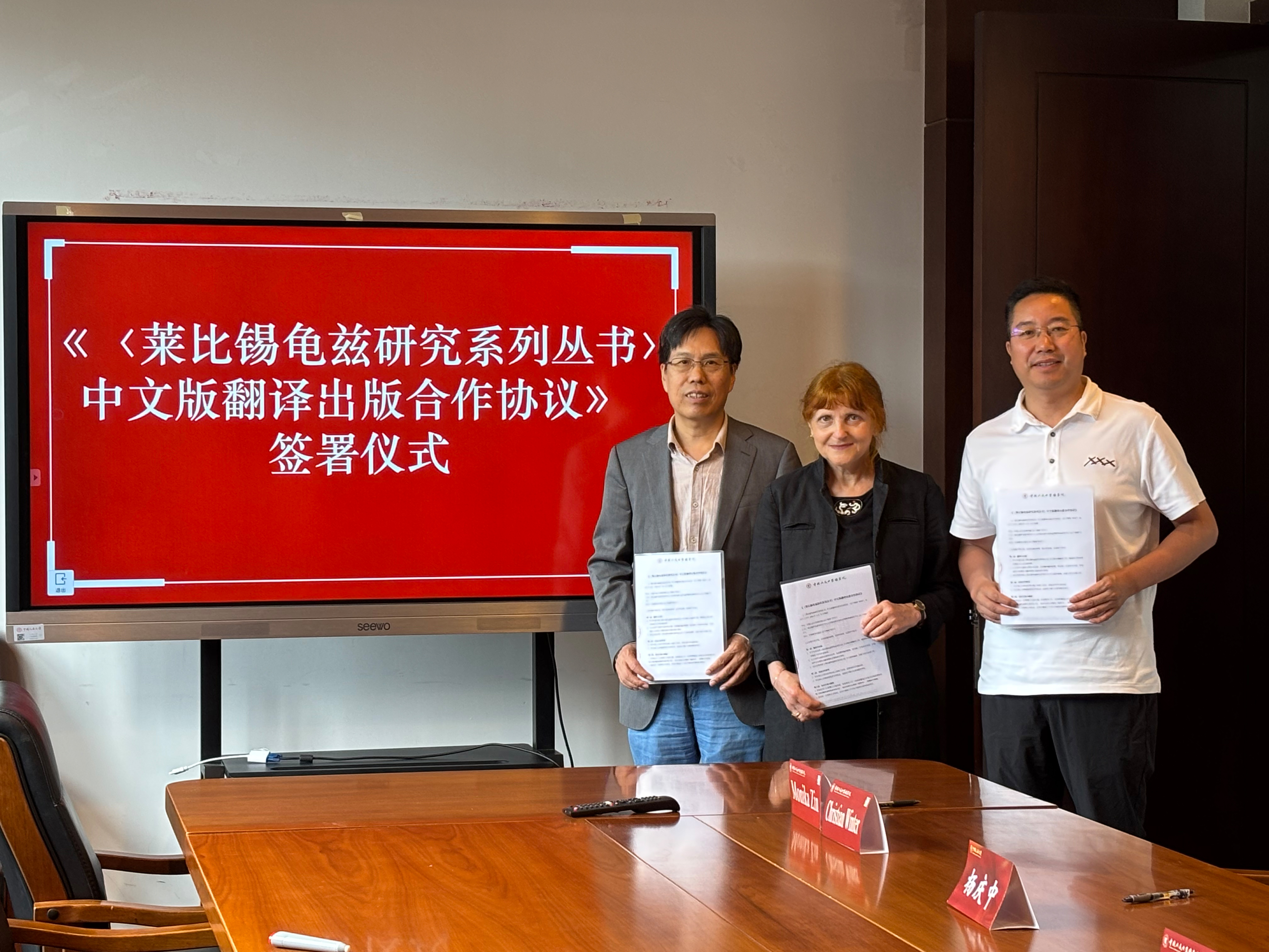 Zwei Männer und eine Frau (Mitte) in einem Konferenzraum halten ein Blatt Papier vor sich, die Kooperationsvereinbarung. Hinter ihnen steht ein großer Flachbildschirm mit weißer chinesischer Schrift auf rotem Hintergrund.