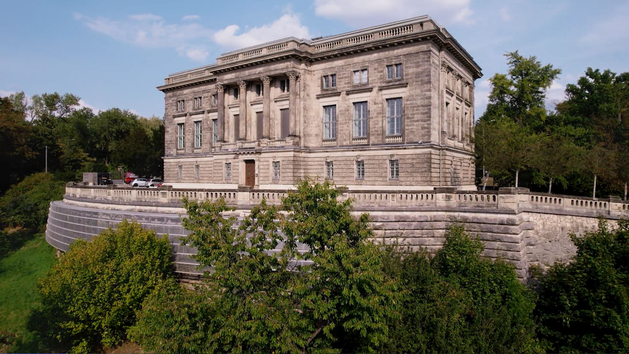 Gebäude mit Säulen und weitläufiger Terasse, umgeben von Bäumen.
