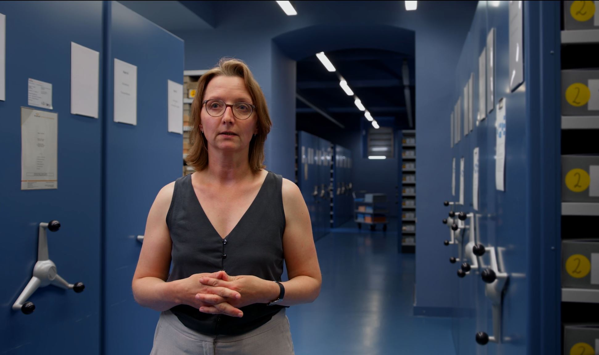 Eine weibliche Person steht vor blauen Rollschränken mit Archivkästen, auf denen Nummern zu sehen sind.