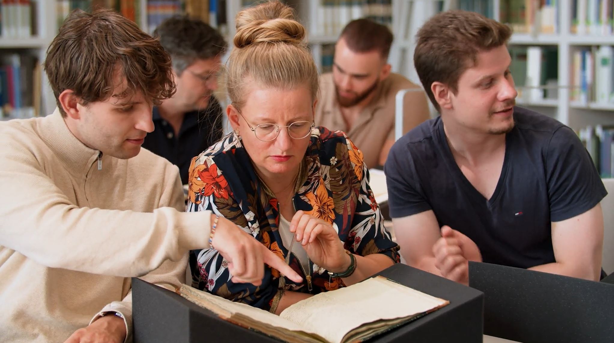 Mehrere Personen sitzen in einem Raum mit Bücherwänden hinter einer aufgeschlagenen alten Handschrift und diskutieren.