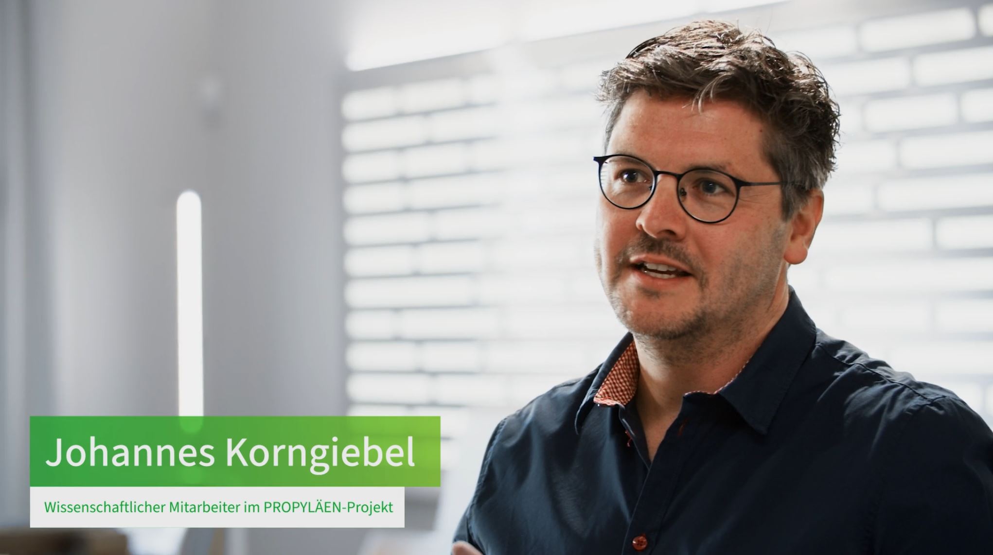Eine männliche Person steht vor einer beleuchteten Wand und spricht in die Kamera. Auf grünem und weißem Untergrund ist unten im Bild zu lesen "Johannes Korngiebel Wissenschaftlicher Mitarbeiter im Propyläen-Projekt"