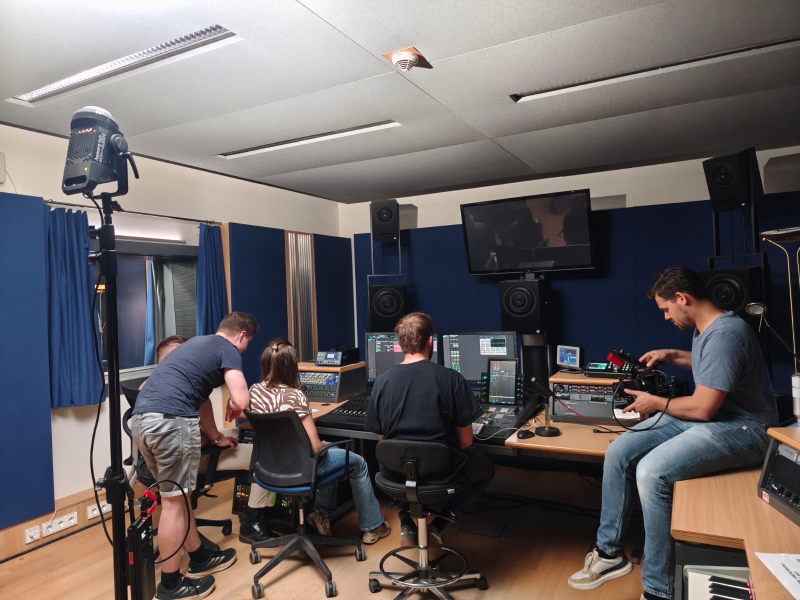 In einem Tonstudio sitzen mehrere Personen vor Reglern und Bildschirmen. Eine Person macht Filmaufnahmen.
