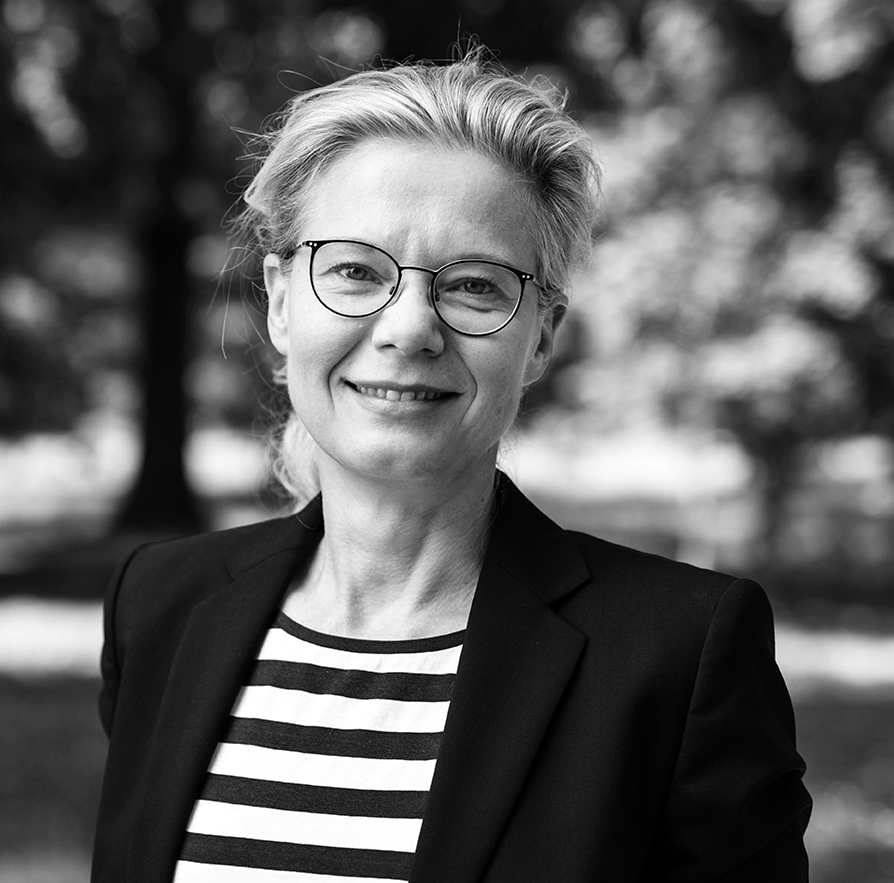 Schwarz-weißes Porträtbild einer lächelnden Frau mit Brille und blonden Haaren. Sie trägt ein gestreiftes Oberteil und Jackett und steht auf einer Wiese vor Bäumen.