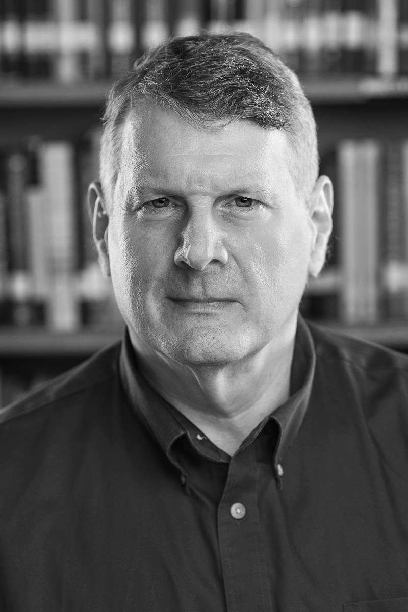 A black-and-white portrait of a man standing in front of a bookshelf