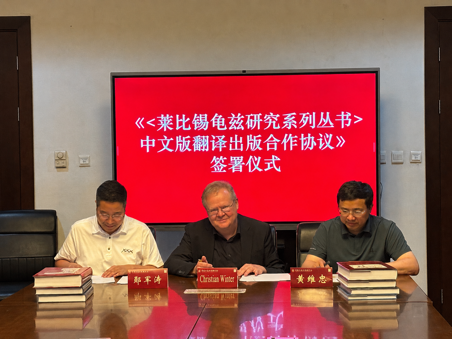 Three men seated at a conferenc table with a red screen and white Chinese writing behind them